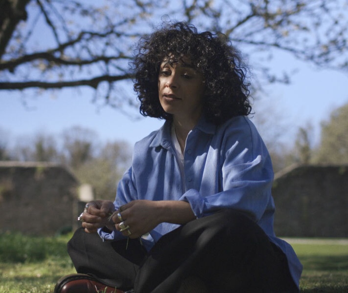 Jade Jordan sits on the grass at Coole Park with a tree and a high wall in the background.