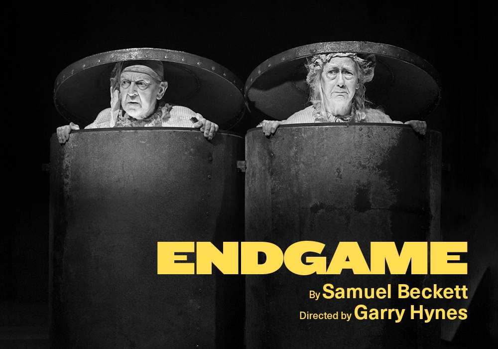 A black-and-white photograph of two people in trash cans with the lids on top of their heads. In the bottom right in red is the show title and names of the writer and director.