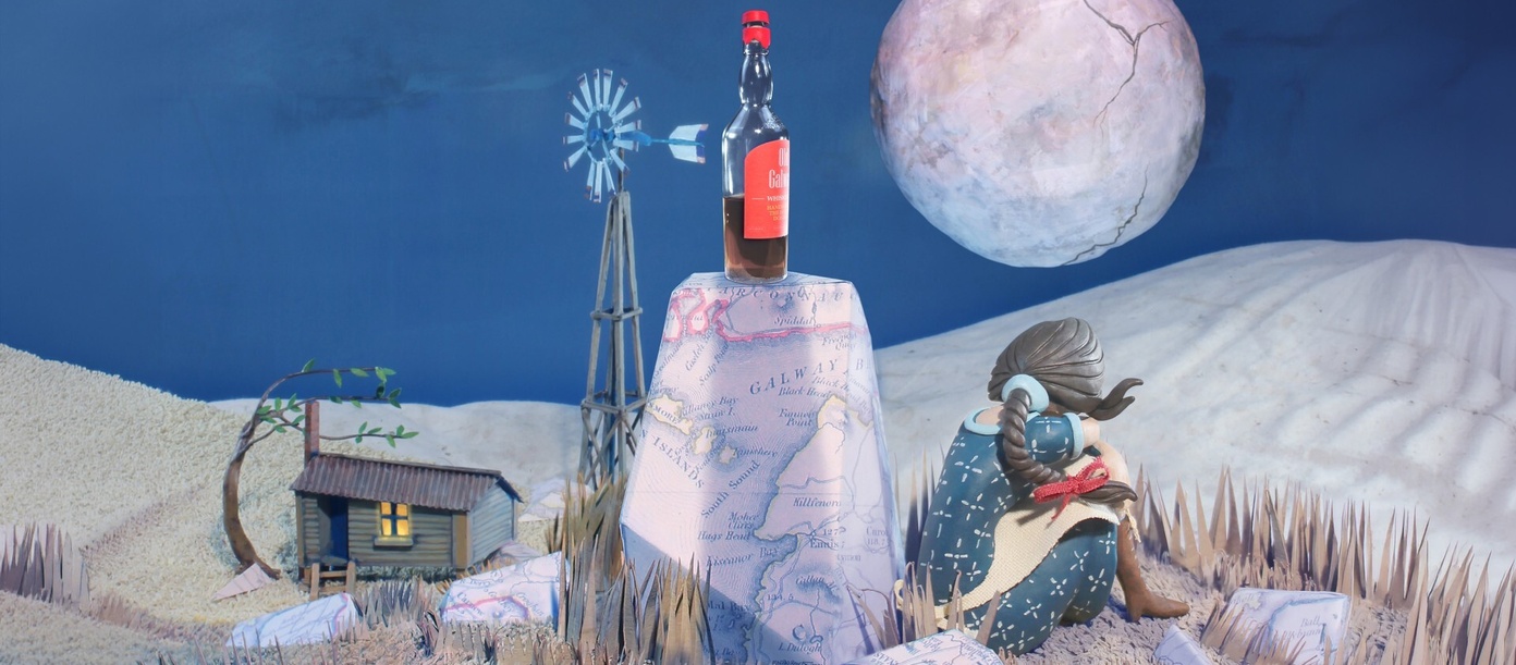 Artwork depicts a nighttime scene, a woman with her back to us sits beside a rock with a bottle of whisky on it. In the background we see a shack, and full moon.
