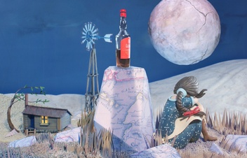 Artwork depicts a nighttime scene, a woman with her back to us sits beside a rock with a bottle of whisky on it. In the background we see a shack, and full moon.