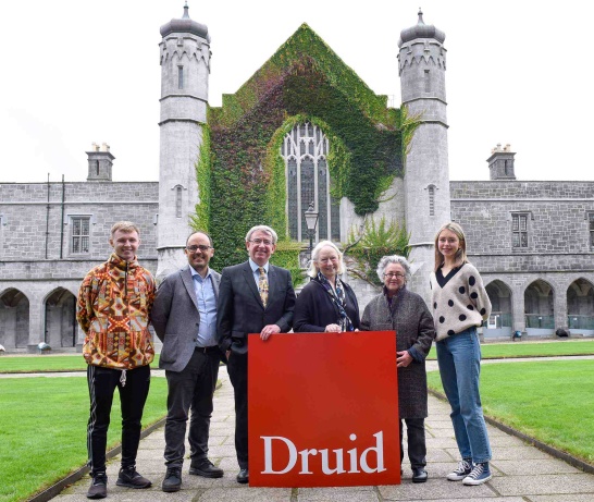 Six people pose in front of a giant Druid logo on campus at the University of Galway.