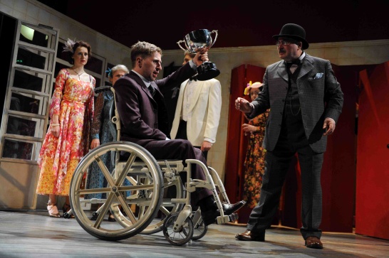 A production photo from The Silver Tassie with actor Aaron Monaghan in the centre seated in a wheelchair and holding a cup aloft.