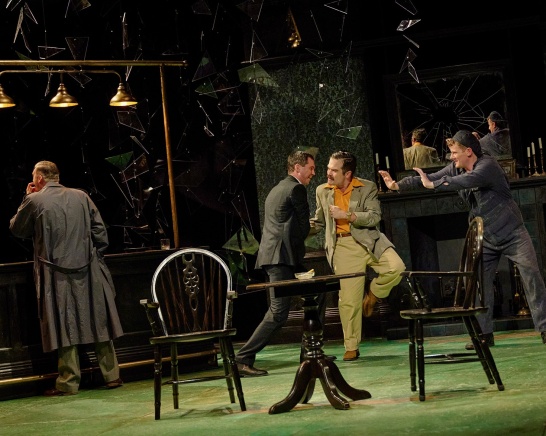 A boisterous scene in the local pub, a group of men play-fight together while another man drinks at the bar.