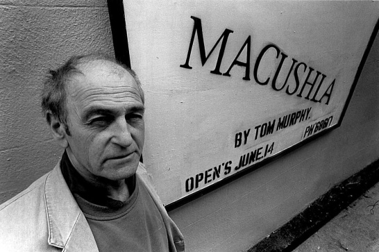 A black-and-white photograph of playwright Tom Murphy outside the Druid Lane Theatre in 1995. There's a large sign on the wall behind him advertising his play The Blue Macushla.