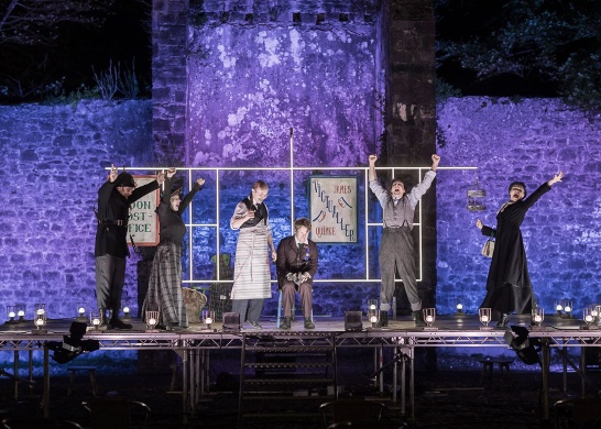The cast of DruidGregory on an outdoor stage in the courtyard of Coole Park with a purple wash of lights against the stone wall behind the stage.