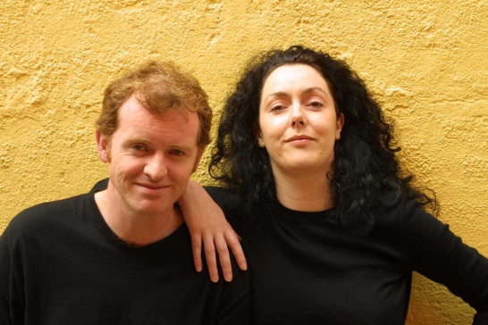 A promotional photo for The Good Father by Christian O'Reilly featuring Aidan Kelly and Derbhle Crotty, both dressed in black and standing in front of a yellow wall.