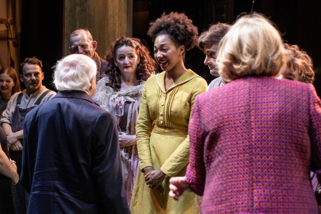 President Higgins with his back to the camera speaks to a group of actors on stage.