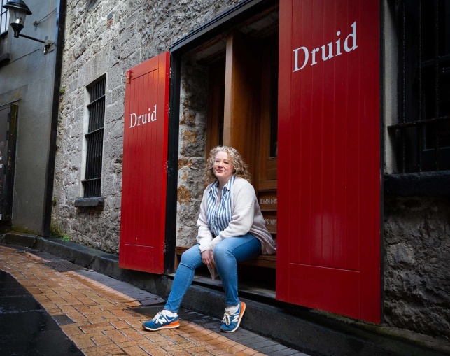 Lianne O'Shea, the 2025 recipient of Druid's Marie Mullen Bursary, seated outside The Mick Lally Theatre on Druid Lane with the red Druid doors open on either side of her.