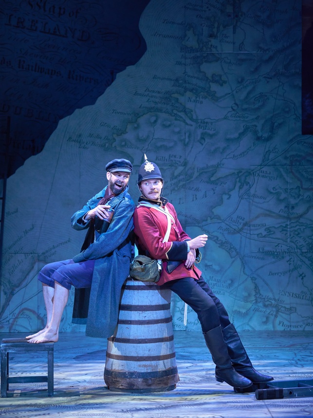 Marty Rea as Robert Ffolliott and Fintan Kinsella as Captain Molineux sitting back to back on a barrel drinking whiskey