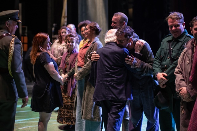 President Higgins with his back to the camera speaks to a group of actors on stage.