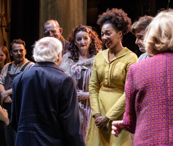 President Higgins with his back to the camera speaks to a group of actors on stage.