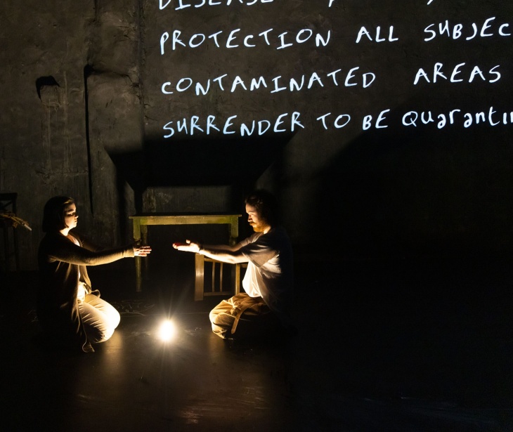 Two people kneel on the floor opposite one another with arms outstretched. Behind on the wall, numerous lines of text are projected in white.