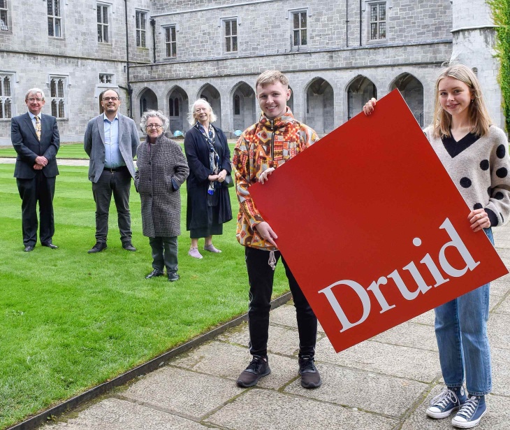 Six people pose in front of a giant Druid logo on campus at the University of Galway.