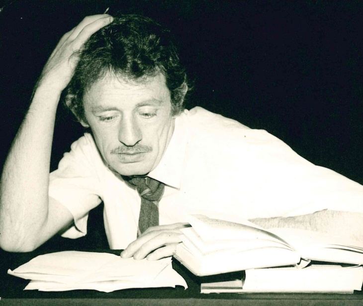 A black-and-white photo of Mick Lally seated at a desk in a shirt and tie with a pile of books on the desk.
