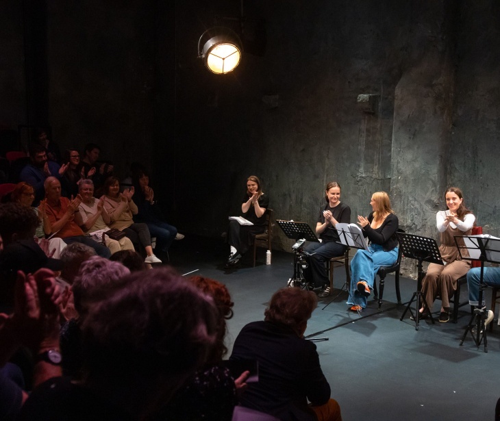 The interior of The Mick Lally Theatre with four people on stage seated with play scripts on stands in front of them, and a full audience applauding.