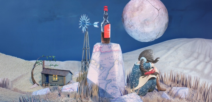 Artwork depicts a nighttime scene, a woman with her back to us sits beside a rock with a bottle of whisky on it. In the background we see a shack, and full moon.