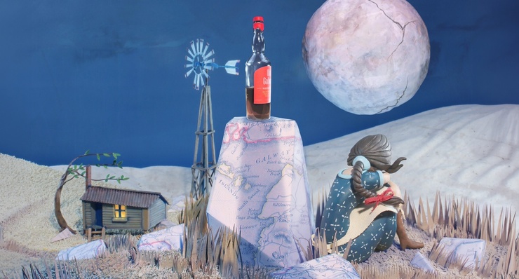 Artwork depicts a nighttime scene, a woman with her back to us sits beside a rock with a bottle of whisky on it. In the background we see a shack, and full moon.