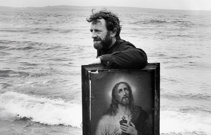 A black-and-white photo of Mick Lally on Inis Meáin with the sea behind him. He's leaning his arm on a large painting of the Sacred Heart which sits on wooden table.