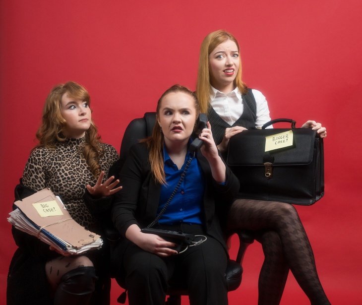 Three actors in costume pose for a promo photo against a red background.